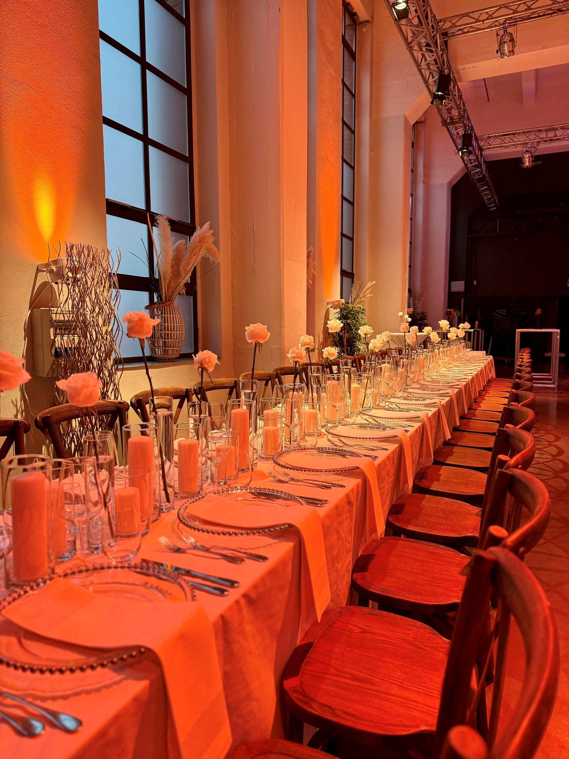 Lange festliche Tafel im Bauwerk Köln mit Holzstühlen, Kerzen und Tischdecken in Orange, warm beleuchtet vor hohen Industriefenstern