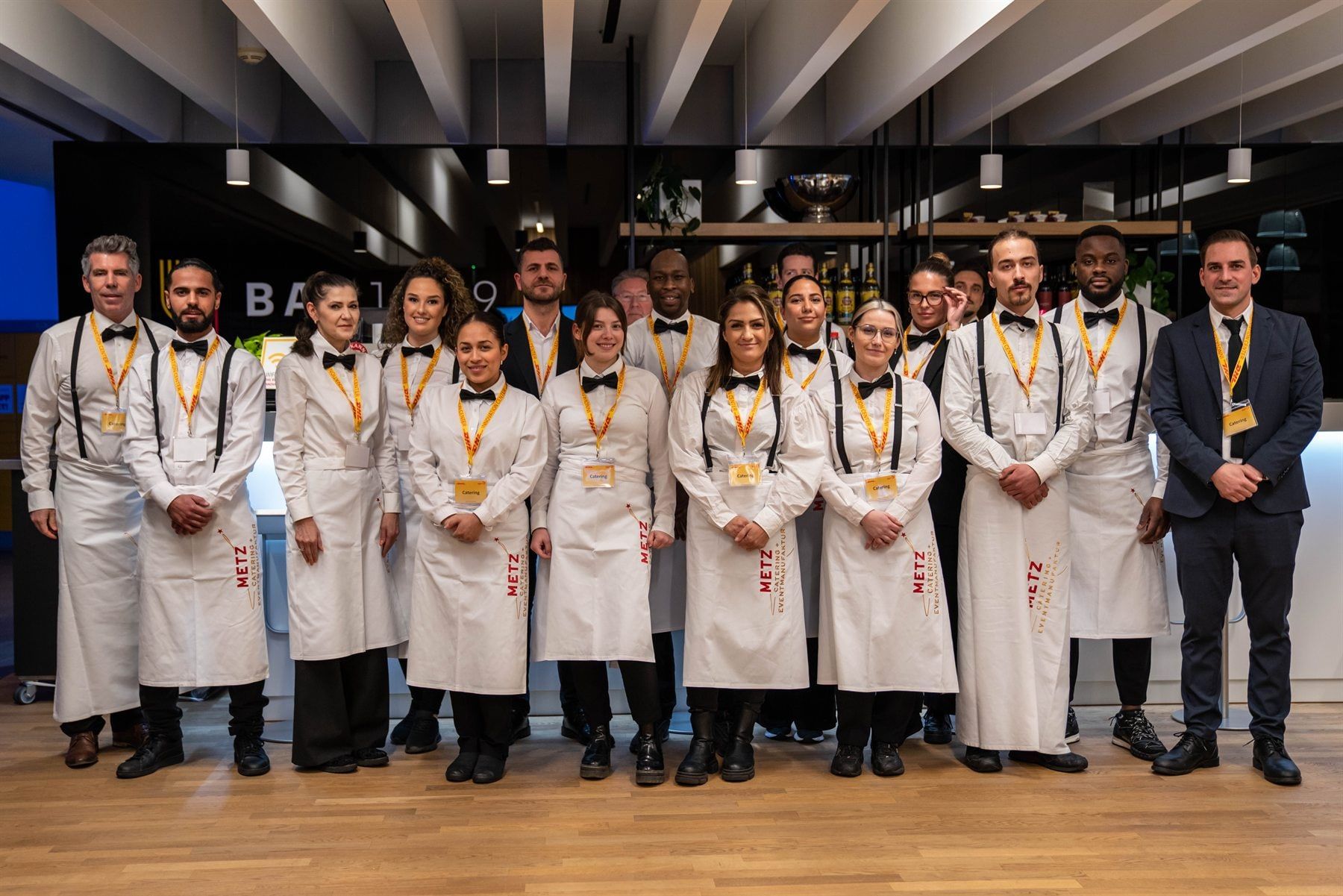 Das Service- und Küchenteam von METZ. in weißen Schürzen und Fliegen, aufgestellt für ein Gruppenfoto vor einem Event
